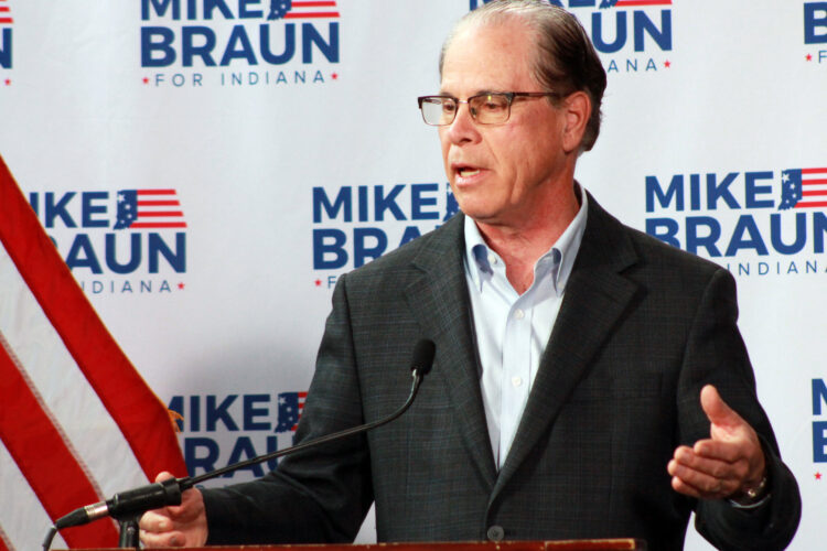 Mike Braun speaks at a lectern. Behind him is a backdrop with his campaign logo dotted across it. Braun is a White man, balding with dark gray hair. He is wearing a dark suit coat and a light-colored shirt.