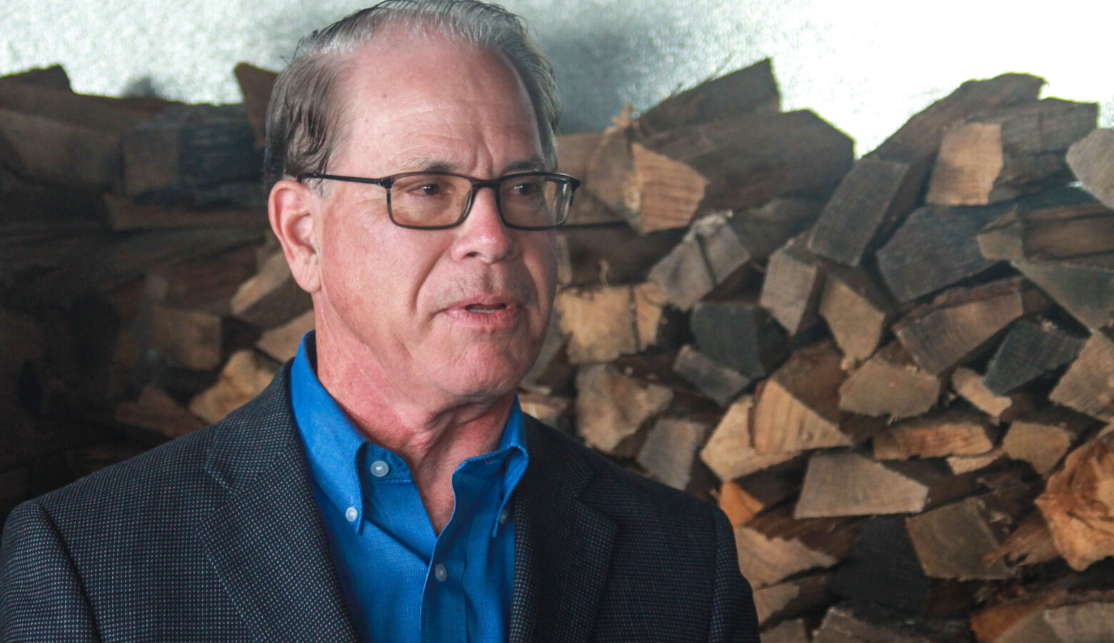 Mike Braun stands in front of a pile of firewood. Braun is a White man with dark, graying hair. He is wearing glasses and a gray sport coat over a blue shirt.