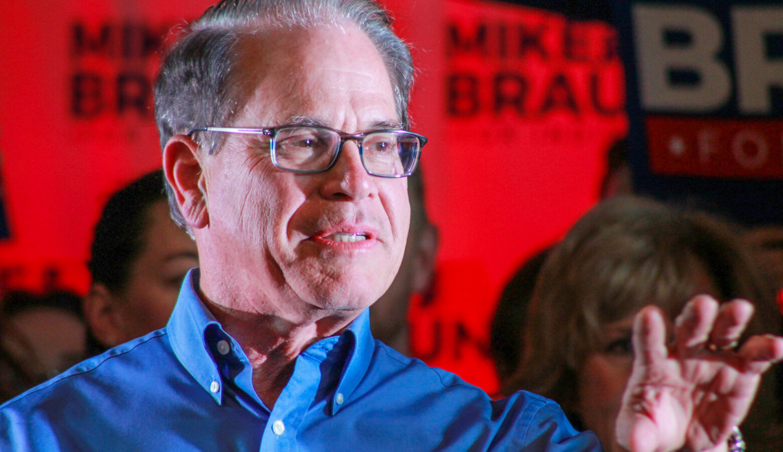 Mike Braun speaks to a crowd with supporters holding signs behind him. Braun is a White man with dark, graying hair. He is wearing glasses and a blue dress shirt.