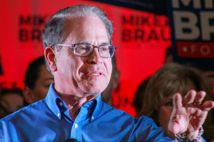 Mike Braun speaks to a crowd with supporters holding signs behind him. Braun is a White man with dark, graying hair. He is wearing glasses and a blue dress shirt.