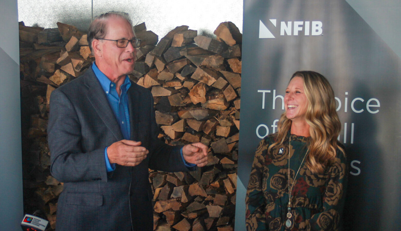 Mike Braun speaks while Natalie Robinson stands beside him. Both are laughing. Braun is a White man with dark, graying hair. He is wearing glasses and a gray jacket over a blue shirt. Robinson is a White woman with blonde hair. She is wearing a green dress with a brown pattern.