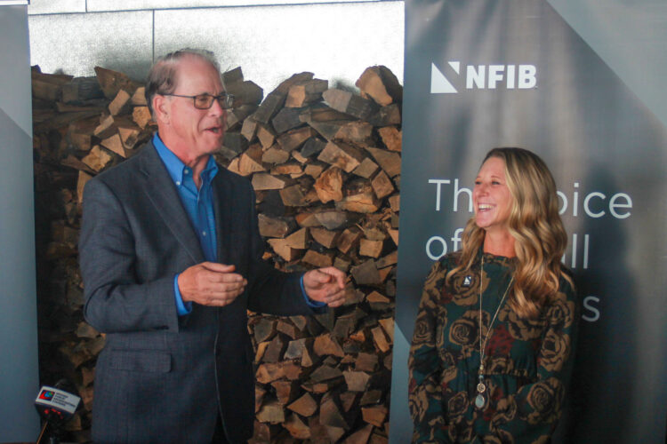 Mike Braun speaks while Natalie Robinson stands beside him. Both are laughing. Braun is a White man with dark, graying hair. He is wearing glasses and a gray jacket over a blue shirt. Robinson is a White woman with blonde hair. She is wearing a green dress with a brown pattern.