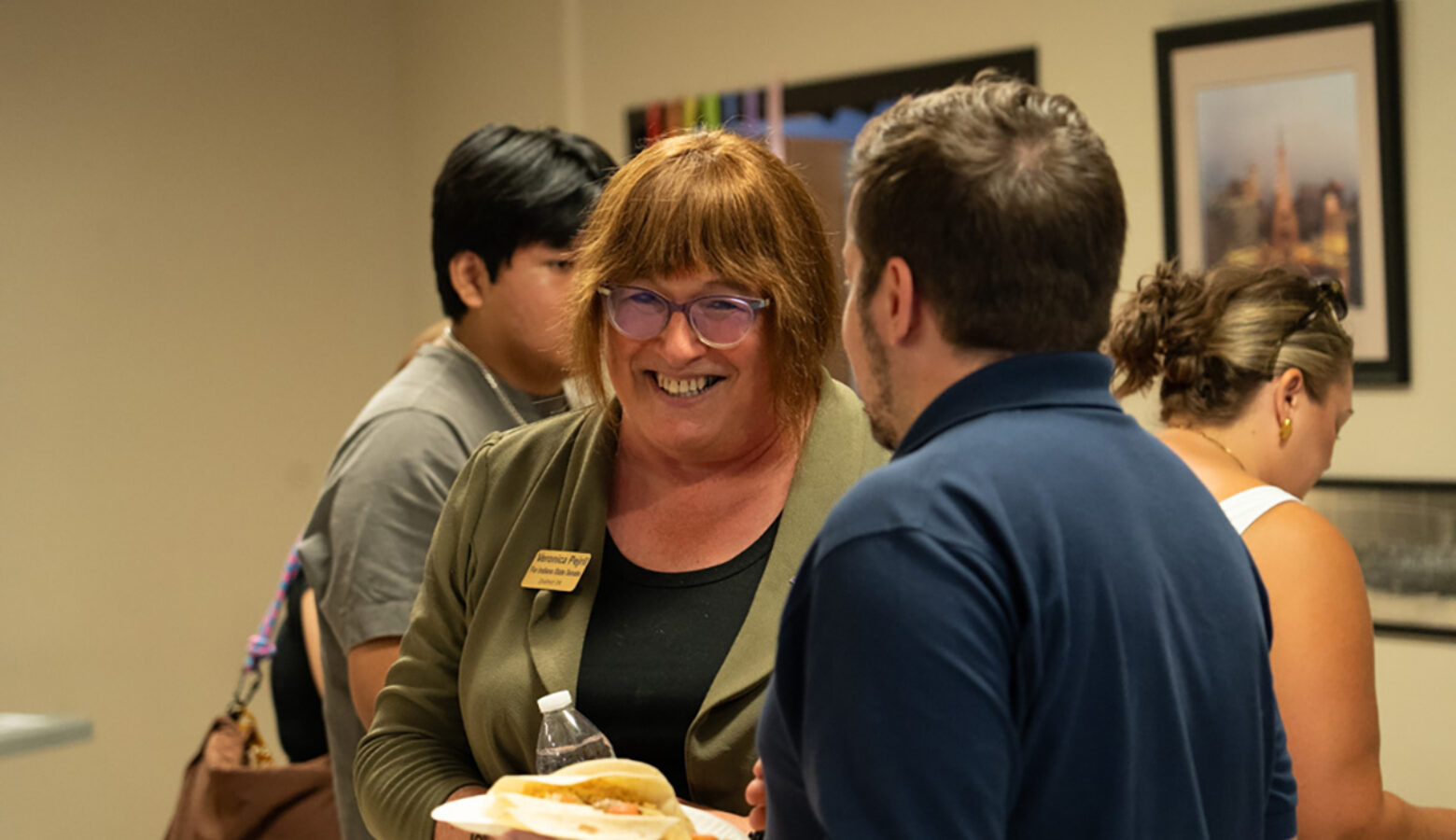 Veronica Pejril wears a dark sage blazer with a name tag, speaking to someone at an event from the Indiana Stonewall Democrats.