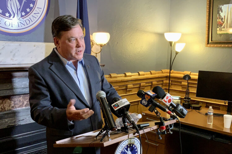 Todd Rokita stands in front of a bank of microphones. Rokita is a White man with dark, graying hair. He is wearing a dark-colored sport coat over a light blue dress shirt.