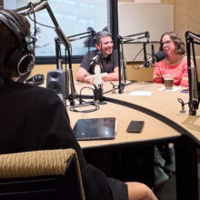 A woman sits facing away from the camera with headphones on. Across the table, a man and a woman smile while talking into microphones.