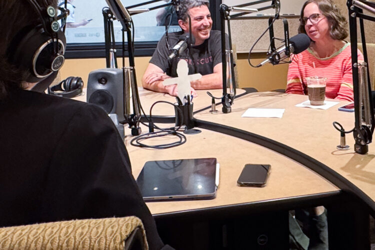 A woman sits facing away from the camera with headphones on. Across the table, a man and a woman smile while talking into microphones.