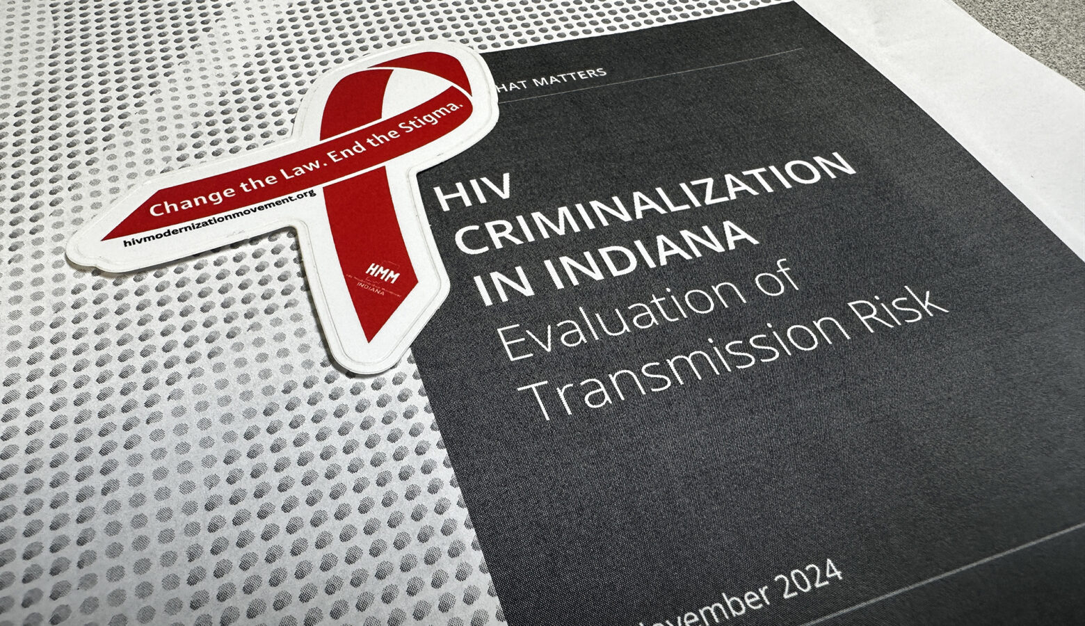 A sticker in the shape of a red ribbon that reads, "Change the Law. End the Stigma." on top of the front page of a report that reads "HIV Criminalization in Indiana: Evaluation of Transmission Risk."