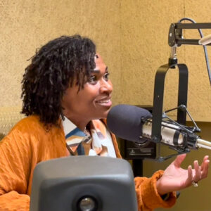 A woman in a yellow sweater sits in front of a microphone smiling an gesticulating.