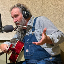 A man in a button up shirt and overalls sits in front of a microphone. He has a guitar in his hands.