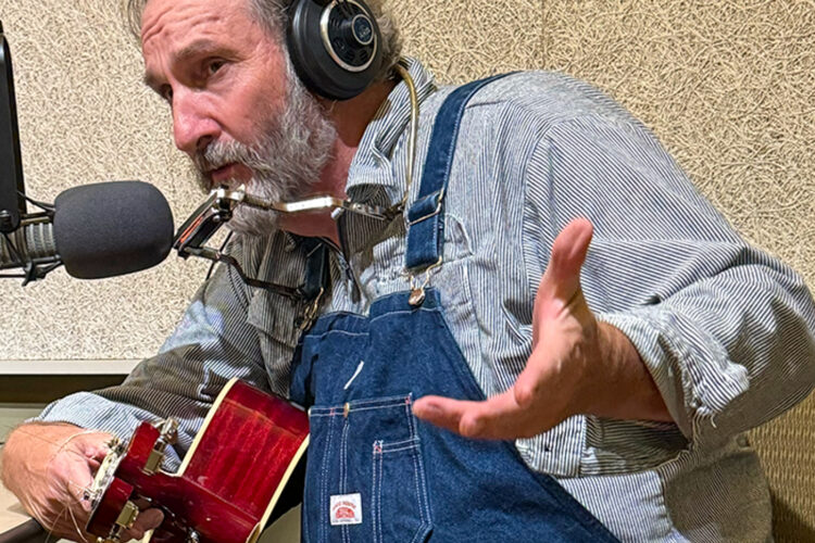 A man in a button up shirt and overalls sits in front of a microphone. He has a guitar in his hands.