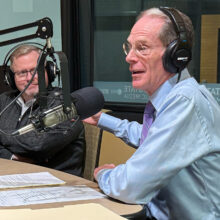 Two men sit at a desk with headphones on, smiling and talking into microphones.