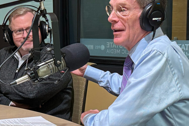 Two men sit at a desk with headphones on, smiling and talking into microphones.