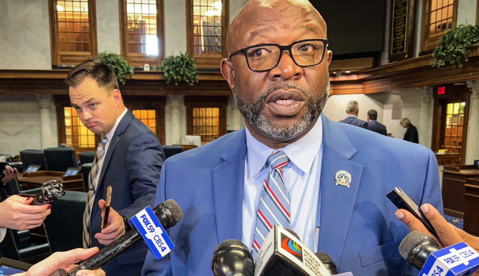 Greg Taylor speaks in front of a bank of microphones. Taylor is a Black man, bald with a dark, graying beard. He is wearing glasses and a suit and tie.