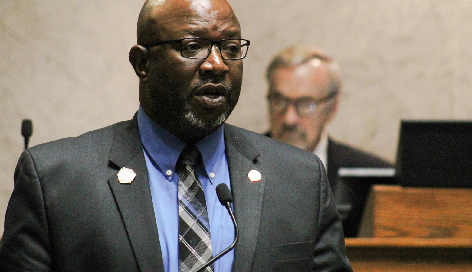 Greg Taylor is a Black man, bald, with a white goatee. Taylor is wearing glasses and a suit and tie.