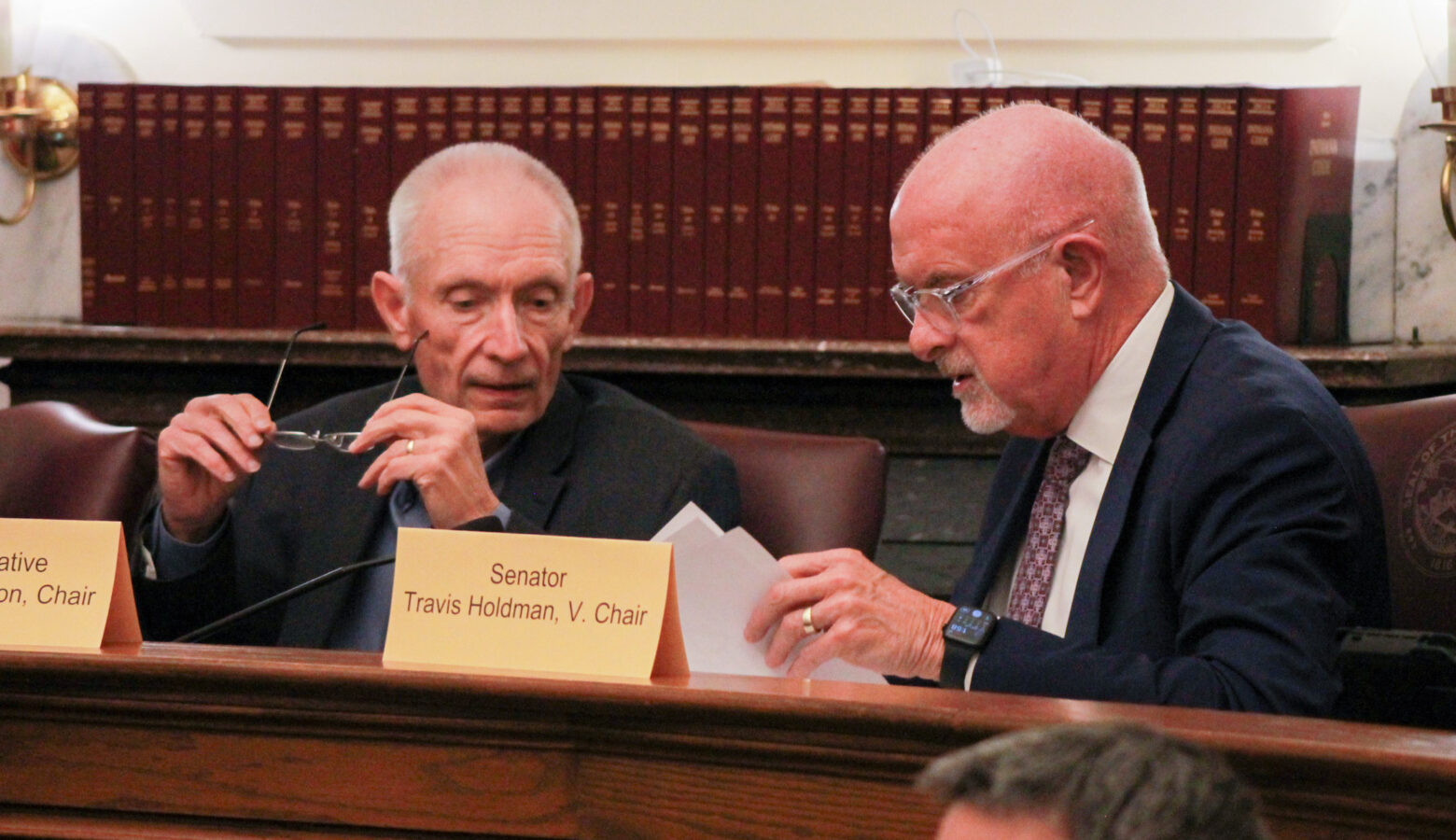 Jeff Thompson and Travis Holdman speak to each other while seated in a committee room. Thompson is a White man with short, white hair. He is wearing a suit and tie. Holdman is a White man, bald, with a white goatee. He is wearing glasses and a suit and tie.