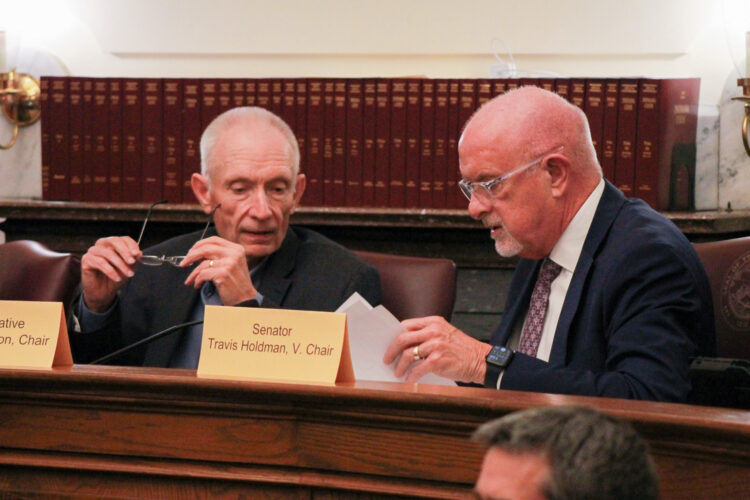 Jeff Thompson and Travis Holdman speak to each other while seated in a committee room. Thompson is a White man with short, white hair. He is wearing a suit and tie. Holdman is a White man, bald, with a white goatee. He is wearing glasses and a suit and tie.