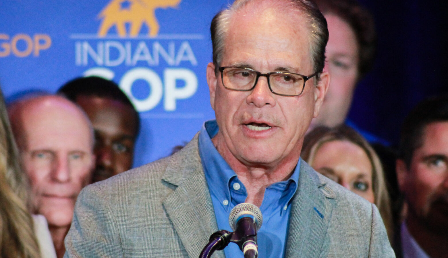 Mike Braun speaks into a microphone on a stage. Braun is a White man, balding, with dark graying hair. He is wearing glasses, a gray suit jacket and blue shirt.