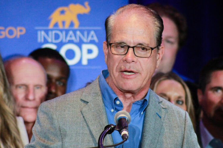 Mike Braun speaks into a microphone on a stage. Braun is a White man, balding, with dark graying hair. He is wearing glasses, a gray suit jacket and blue shirt.