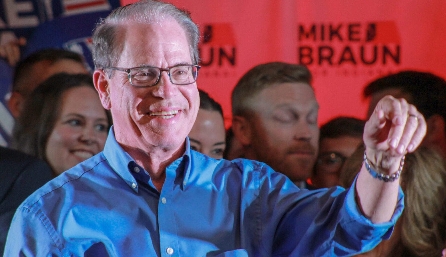 Mike Braun stands on a stage, gesturing while supporters stand behind him. Braun is a White man, balding with gray hair. He is wearing glasses and a blue shirt.