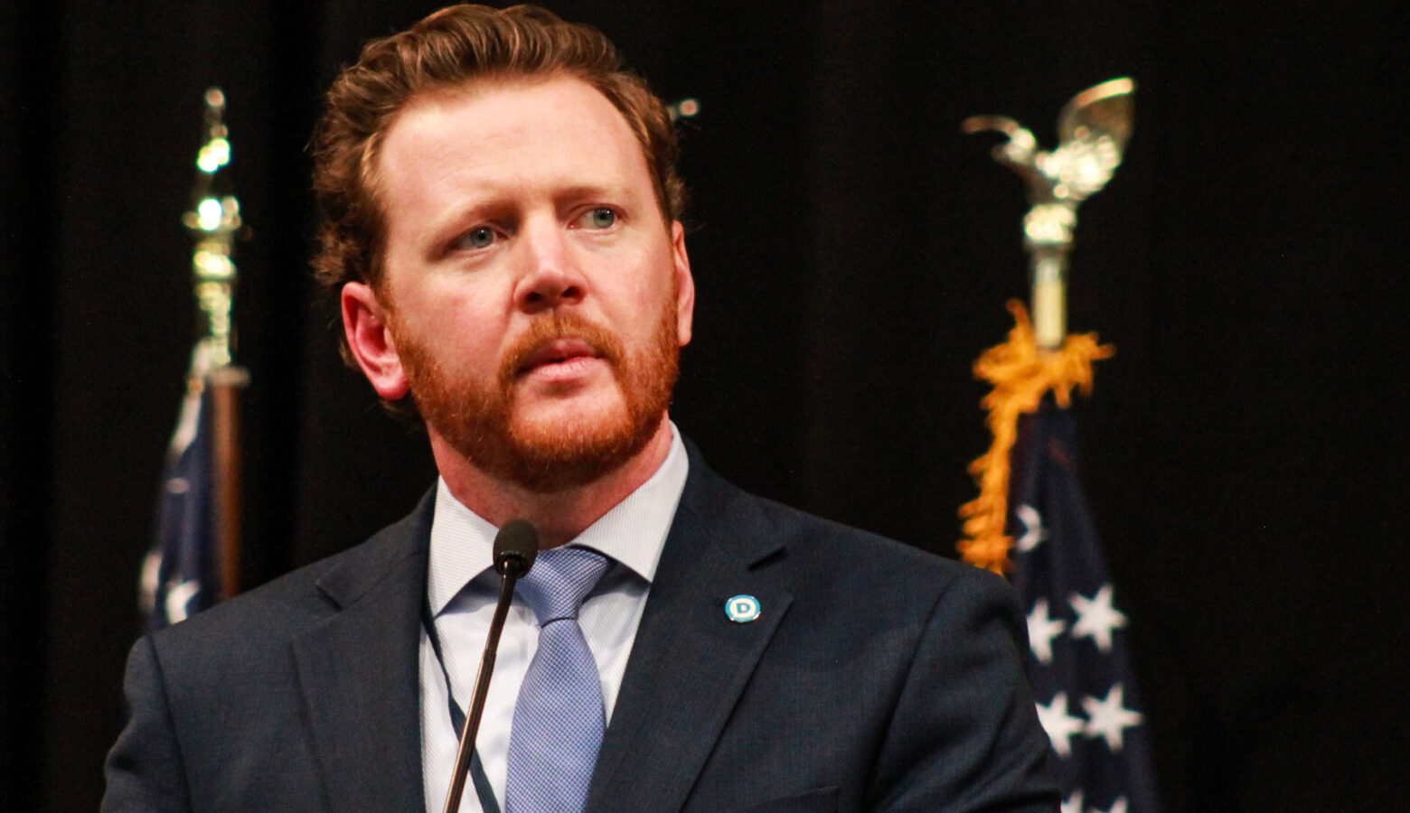 Mike Schmuhl stands at a lectern, with two American flags standing behind him. Schmuhl is a White man with red hair and beard. He is wearing a suit and tie.