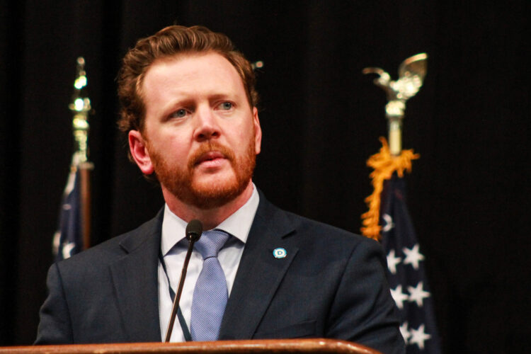 Mike Schmuhl stands at a lectern, with two American flags standing behind him. Schmuhl is a White man with red hair and beard. He is wearing a suit and tie.