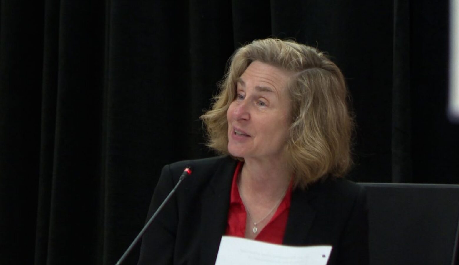 Pamela Whitten sits, speaking into a microphone. Whitten is a White woman with blonde hair. She is wearing a black jacket and a red shirt.