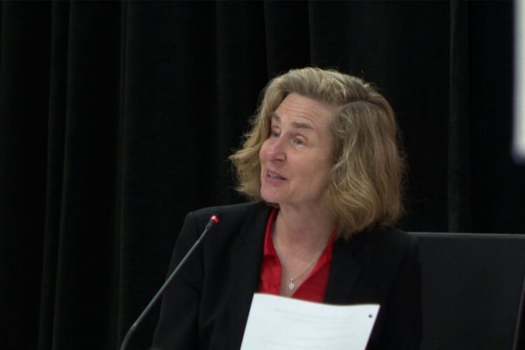 Pamela Whitten sits, speaking into a microphone. Whitten is a White woman with blonde hair. She is wearing a black jacket and a red shirt.