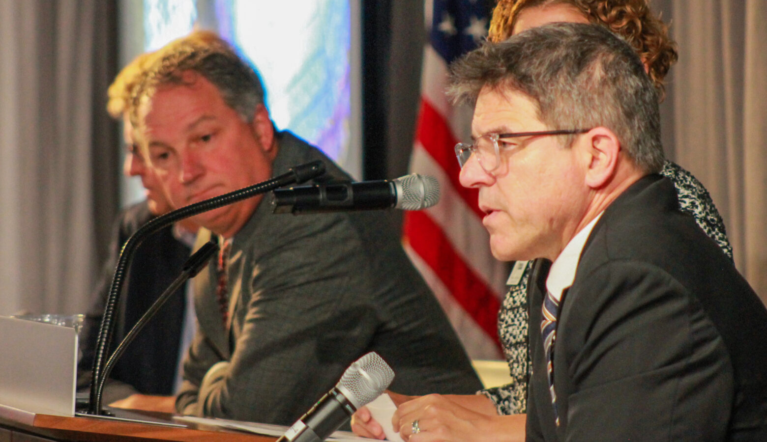 Rodric Bray sits on a stage and talks into a microphone while Todd Huston, also seated, looks on in the background. Bray is a White man with dark, graying hair. He is wearing glasses and a suit and tie. Huston is a White man with gray hair. He is wearing a suit and tie.