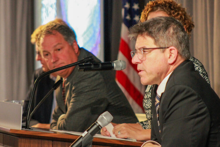 Rodric Bray sits on a stage and talks into a microphone while Todd Huston, also seated, looks on in the background. Bray is a White man with dark, graying hair. He is wearing glasses and a suit and tie. Huston is a White man with gray hair. He is wearing a suit and tie.