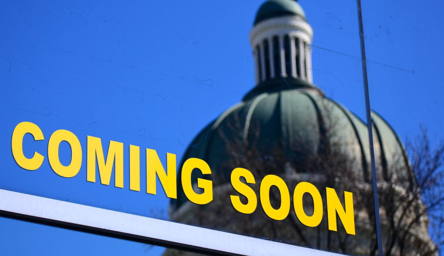 The Indiana Statehouse is seen in a reflection from windows in a building across the state. A graphic on the window reads "Coming Soon"