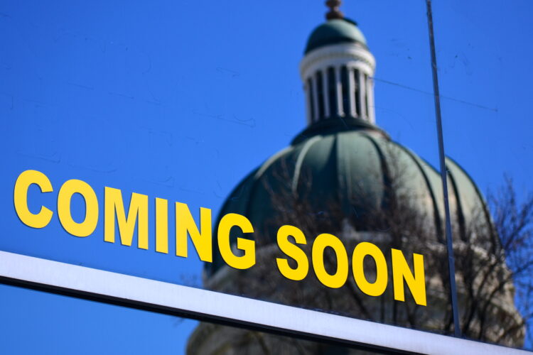 The Indiana Statehouse is seen in a reflection from windows in a building across the state. A graphic on the window reads "Coming Soon"