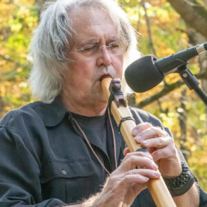 A man with shoulder-length gray hair and glasses plays a long wooden flute. His eyes are closed in concentration.