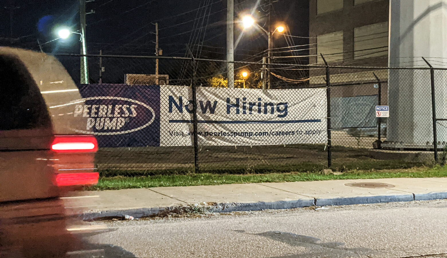 A car drives by a canvas sign fixed to a wire fence that reads "Now Hiring"