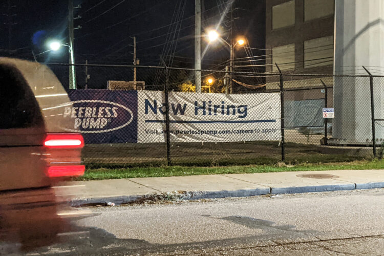 A car drives by a canvas sign fixed to a wire fence that reads "Now Hiring"