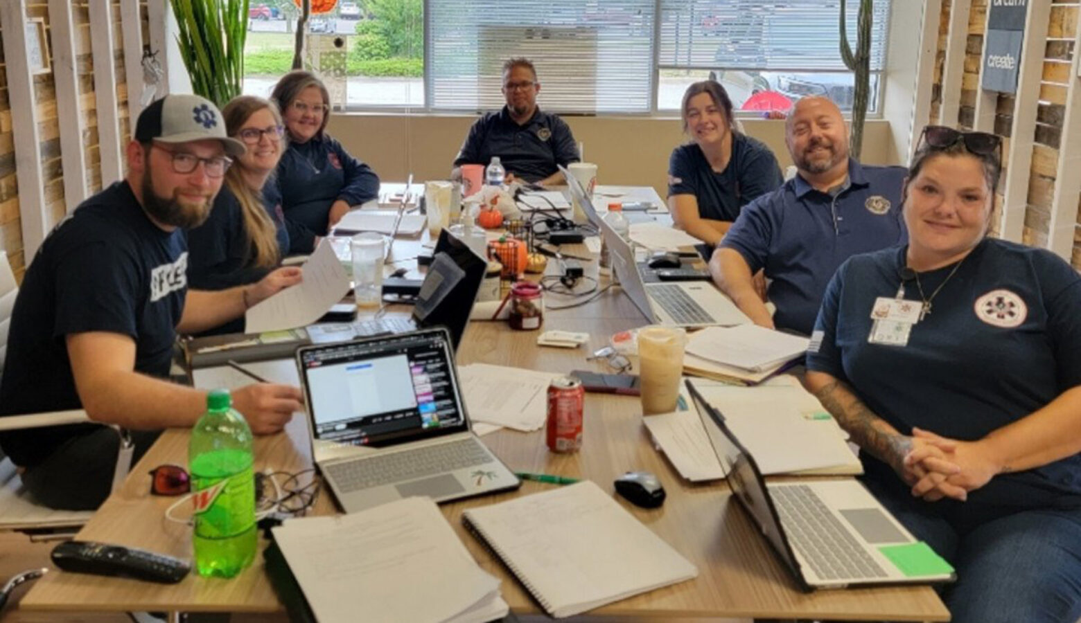 A group of people from Phoenix Paramedic Solutions sit around a table, posing for a camera. The table is covered in papers and laptops.