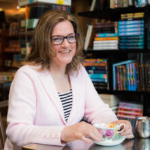 A woman sit smiling at a table with a wall of books behind her. She's holding a teacup on a saucer.