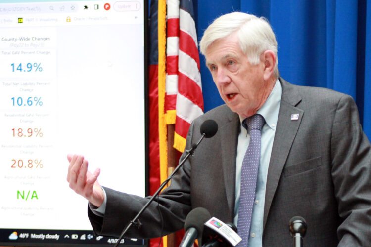 Ed DeLaney is a White man, with white hair. He is speaking into a microphone at a podium, wearing a grey suit. The American Flag is in behind his shoulder.