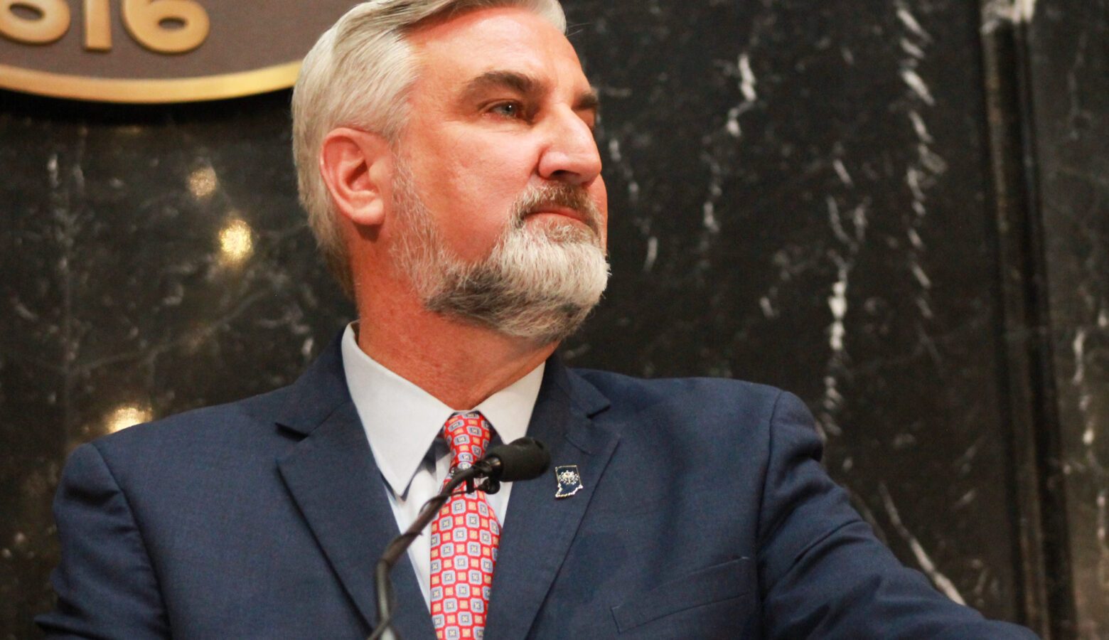 Eric Holcomb stands in front of a microphone in the Indiana House. Holcomb is a White man with white and gray hair and beard. He is wearing a blue suit.