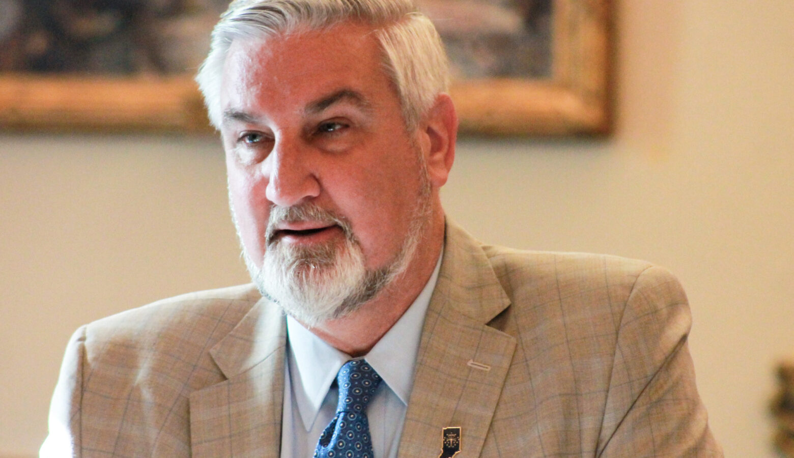 Eric Holcomb is a White man with white hair and beard. He is wearing a tan suit and blue tie.