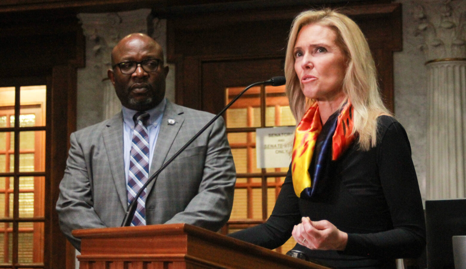 Shelli Yoder speaks into a microphone at a lectern on the Senate floor while Greg Taylor looks on. Yoder is a White woman with blonde hair. She is wearing a black top and a red and yellow scarf. Taylor is a Black man, bald with a goatee. He is wearing glasses and a gray suit.