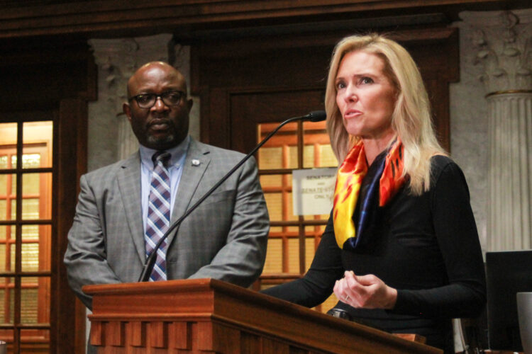 Shelli Yoder speaks into a microphone at a lectern on the Senate floor while Greg Taylor looks on. Yoder is a White woman with blonde hair. She is wearing a black top and a red and yellow scarf. Taylor is a Black man, bald with a goatee. He is wearing glasses and a gray suit.