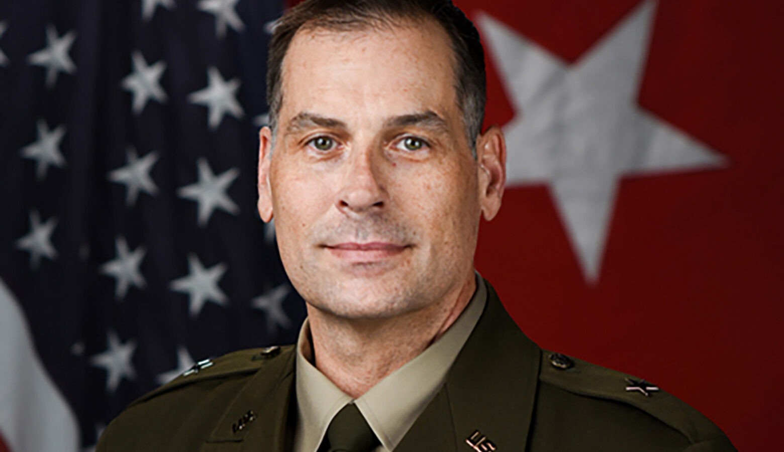 A portrait photograph of Larry Muennich. He is a White man with dark, slightly graying hair. He is standing in front of two flags and wearing his military uniform.