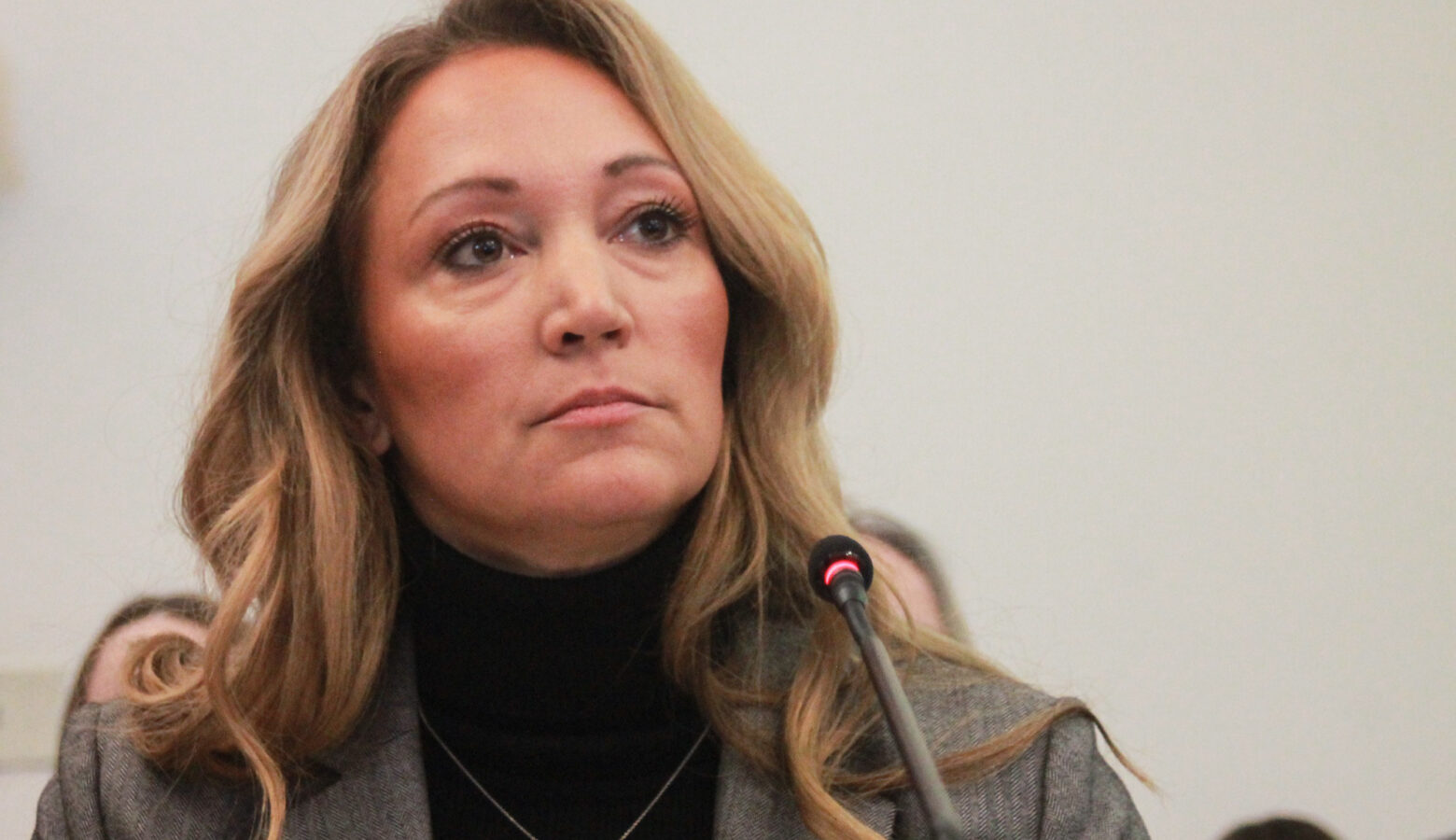 Lindsay Weaver sits in front of a microphone. Weaver is a White woman with dark blonde hair. She is wearing a gray blazer over a black turtleneck.