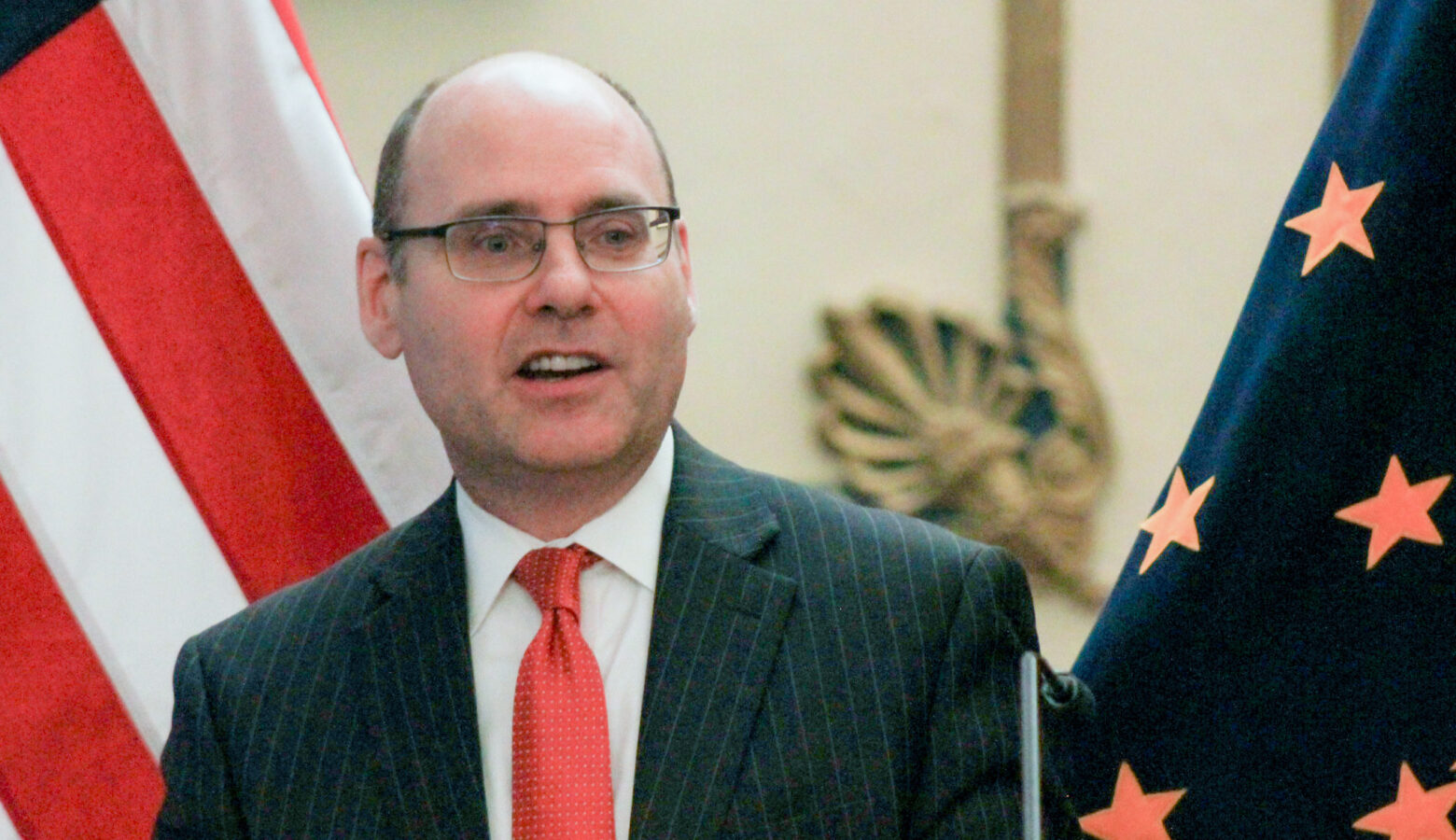 Stephen Scheele stands in front of the American flag and the Indiana flag as he speaks. Scheele is a White man, balding with dark hair. He is wearing glasses and a suit and tie.