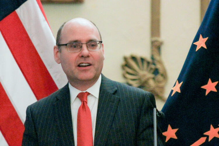 Stephen Scheele stands in front of the American flag and the Indiana flag as he speaks. Scheele is a White man, balding with dark hair. He is wearing glasses and a suit and tie.