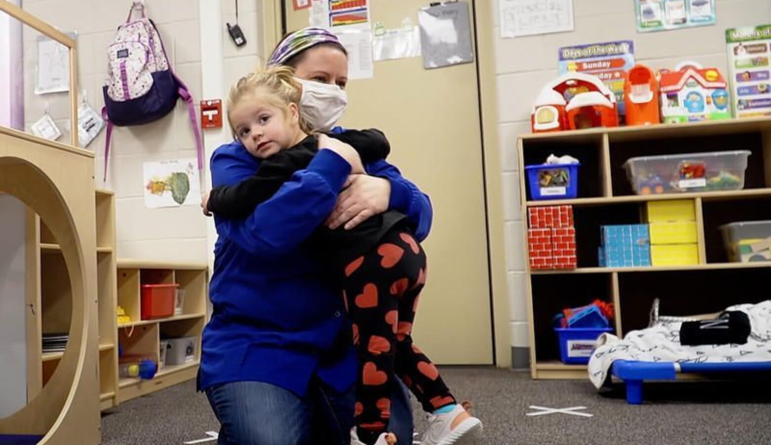 A child receives a hug from a child care worker wearing a mask.