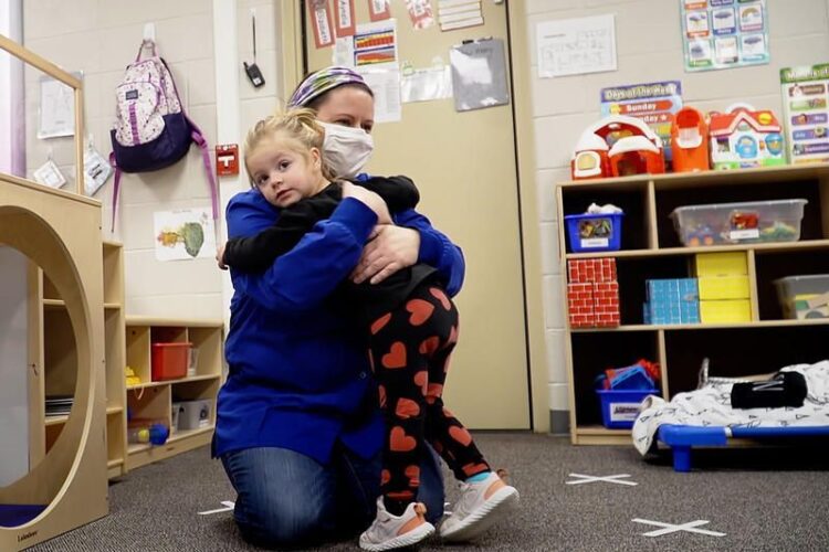 A child receives a hug from a child care worker wearing a mask.