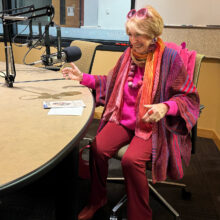 A woman in a bright outfit laughs sitting by a microphone in a studio.