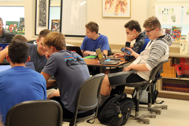 A group of high school students sit in a classroom, working on laptops.