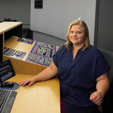 A woman with blond hair and a smile sits at a console with buttons, knobs, and screens.